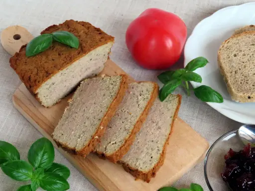 Pokrojony pasztet mieszany drobiowo-wieprzowy na desce, obok pomidor, chleb i konfitura.