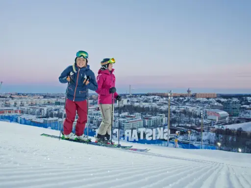 Två skidåkare njuter av utsikten över Hammarbybacken och staden i skymningen.