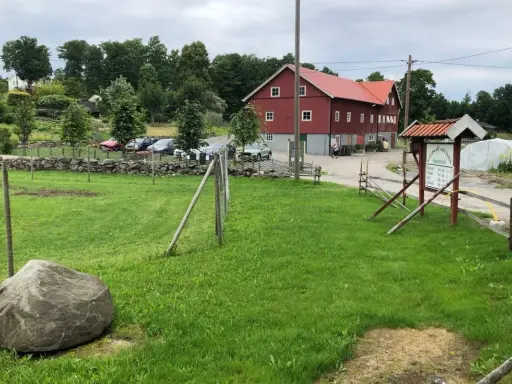 Grönskande gräsmatta med en stor sten framför en röd ladugård på Sammels lantgård. En skylt visar vägen till gårdskafé och butik.