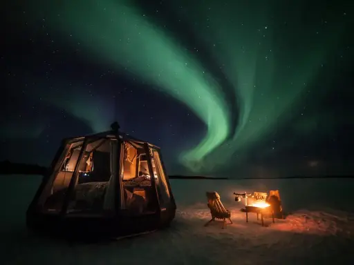En glaskupol för camping i Norrland lyser upp natten under ett dansande norrsken. En eld sprakar vid sidan om.