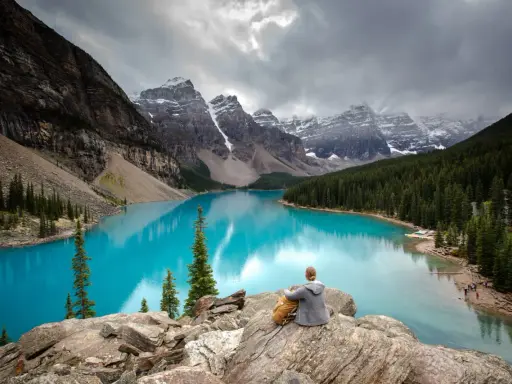 Person sitzt auf Felsen und blickt auf den türkisfarbenen Moränen-See im Banff National Park, Kanada. Majestätische Berge und Nadelwälder umgeben das Wasser.