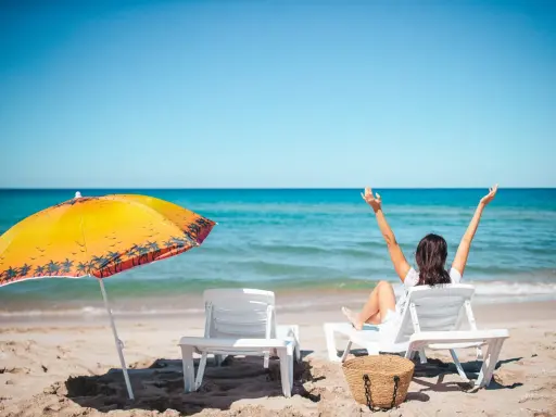 Kobieta cieszy się urlopem wypoczynkowym na plaży, z rękami w górze. Obok leżaki i parasol.