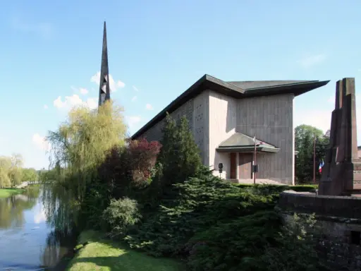 L'église Saint-Rémy de Baccarat, avec sa flèche élancée, se dresse près d'une rivière bordée d'arbres verdoyants.