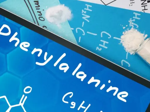 Na ekranie tabletu nazwa "Phenylalanine" i wzór chemiczny. Obok proszek fenyloalaniny w probówce i na kartce.
