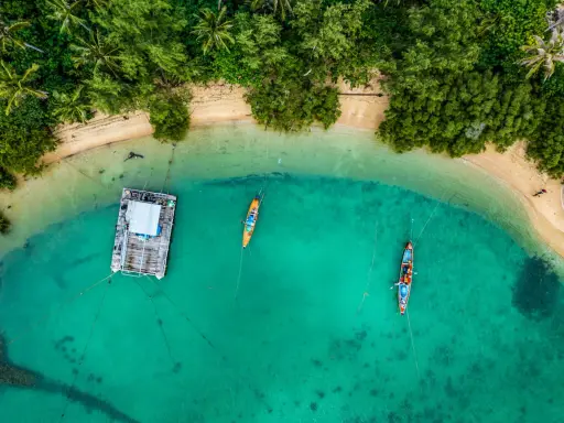 Drohne über Thailand: Ein Blick von oben auf eine schwimmende Plattform und zwei Boote im türkisfarbenen Meer, umgeben von üppiger Vegetation und Sandstrand.