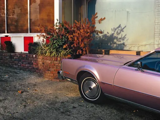 Un'auto rosa pastello, un classico stile William Eggleston, parcheggiata vicino a un muro di mattoni e un edificio con pannelli rossi e bianchi.