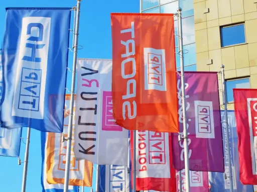 Kolorowe flagi z napisem "TVP w likwidacji" powiewają przed nowoczesnym budynkiem.