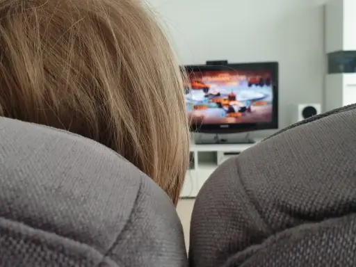 Niemowlę z blond włosami ogląda bajkę na telewizorze w tle, siedząc na kanapie.