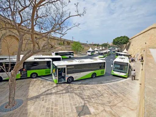 Malta autobusy z lotniska czekają na pasażerów. Wiele zielono-białych pojazdów stoi w rzędzie przy zabytkowych murach.