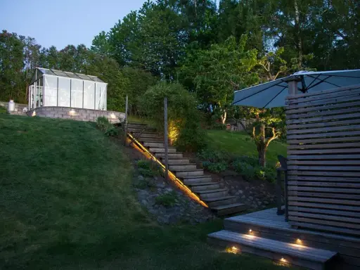 Vacker altan med decklights som lyser upp trappan och trädgården. Ett växthus syns i bakgrunden.