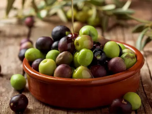 Aceite de arbequina vertiéndose sobre aceitunas verdes y moradas en un cuenco de barro. Ideal para ensaladas y aliños.