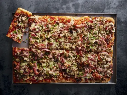 Prostokątna pizza 4 sery z pieczarkami, szynką i oliwkami, pokrojona na kwadraty, z jednym kawałkiem odłożonym na bok.