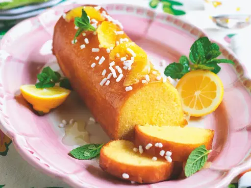 Deliziosa torta al limone glassata, decorata con fette d'arancia, zuccherini e menta. Servita su un piatto rosa con spicchi di limone.