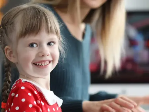 Uśmiechnięta dziewczynka w czerwonej sukience w groszki, z warkoczykami, uczy się kindersztuby od mamy.