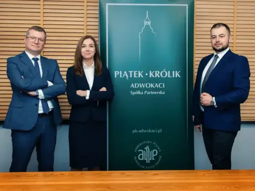 Trzy osoby w garniturach stoją przed zielonym banerem kancelarii "Piątek • Królik Adwokaci".