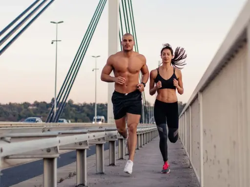 Ein Mann und eine Frau joggen auf einer Brücke. Beide haben sichtbare bauchmuskeln, ein Zeichen für ihr intensives Training.