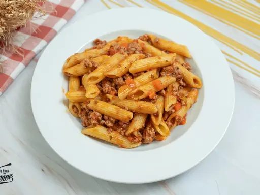Plato de macarrones con carne picada y salsa de tomate, decorado con un paño a cuadros y espaguetis secos al fondo.