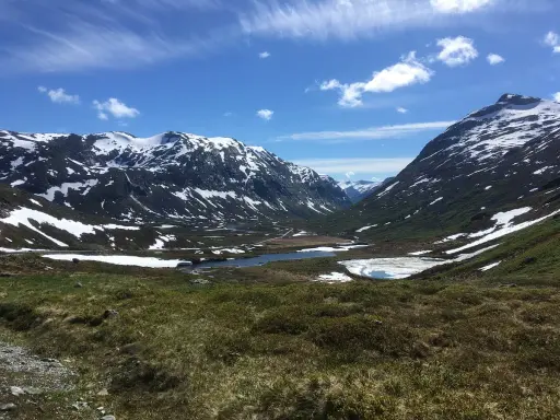 Vacker dalgång med snöklädda berg, en sjö och grönskande sluttningar. En väg slingrar sig fram, kanske en del av trollväggen.