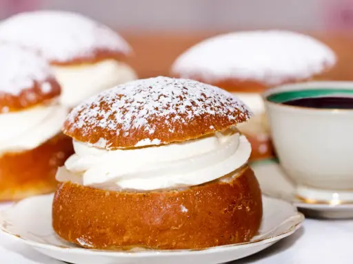Semlor, en fest vid fastan, pudrade med florsocker och fyllda med grädde. En kopp kaffe i bakgrunden.