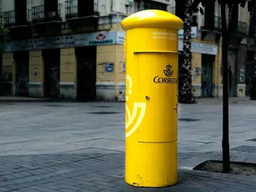 Jasnożółta skrzynka pocztowa Correos, symbol poczty Hiszpanii, stoi na brukowanym placu przed budynkami.