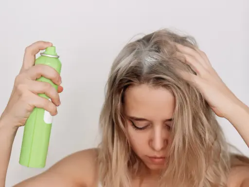 Frau sprüht sich Haarspray auf die Kopfhaut, was die Haare austrocknen kann.