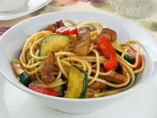 Un plato de espaguetis con pimientos rojos y calabacín salteados, con trozos de carne.