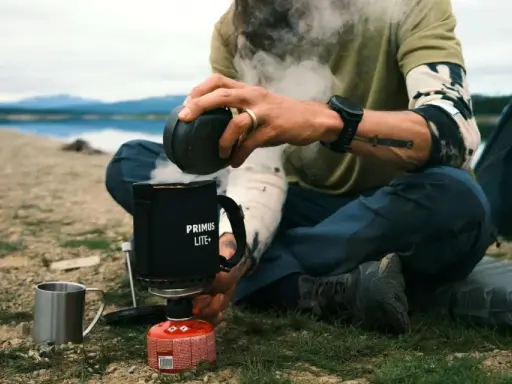 En optimus hiker njuter av en varm dryck vid en sjö. Ånga stiger från koppen medan personen sitter bredvid ett tält.