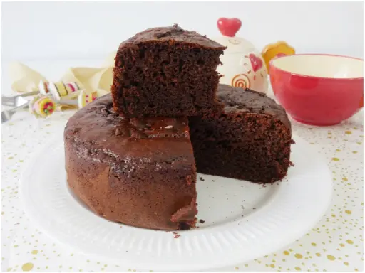 Una fetta di torta yogurt e cioccolato, soffice e golosa, è appoggiata su una torta intera. Sullo sfondo, una tazza rossa e un barattolo decorato.