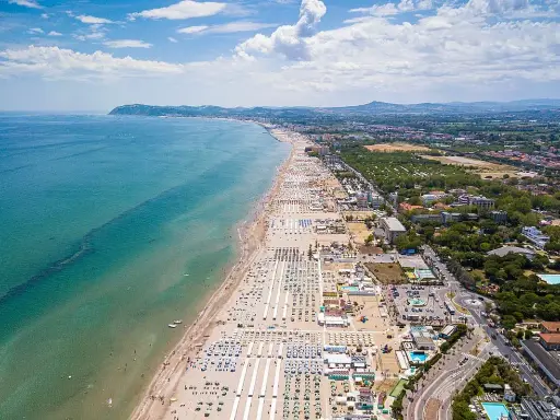 Vista aerea della Riviera Romagnola con spiaggia affollata, mare azzurro e verdeggiante entroterra.