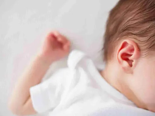 Un neonato dorme troppo, con la testolina appoggiata su un morbido lettino bianco. Si intravede un orecchio rosa e una manina.