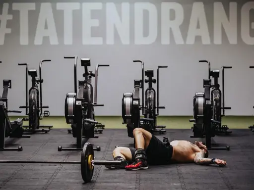 Mann liegt erschöpft auf dem Boden neben einer Hantelstange, umgeben von Trainingsgeräten. Ein Zeichen an der Wand verkündet "#TATENDRANG" – ein Symbol für den Wiedereinstieg in den **Sport nach Corona**.