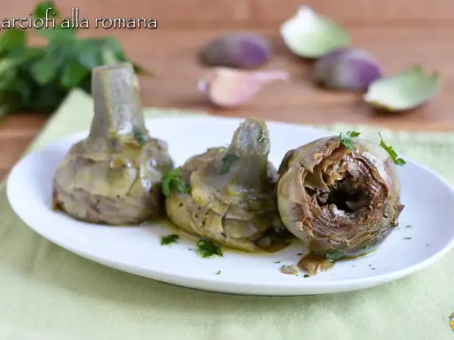 Tre carciofi alla romana cotti, serviti su un piatto bianco con prezzemolo tritato. Sullo sfondo, spicchi d'aglio e foglie di carciofo.