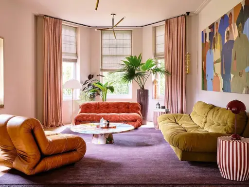Comment amenager un salon : un fauteuil orange, un canapé rouille, un canapé moutarde, une table basse colorée, un tapis violet et une grande peinture.