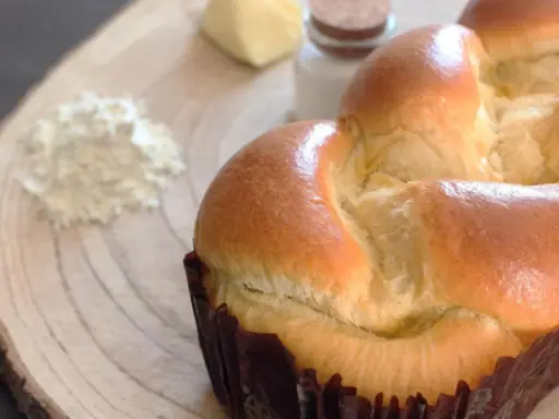 Un pan brioche dorado y esponjoso, listo para ser disfrutado. A su lado, ingredientes para su elaboración: harina, mantequilla y sal.