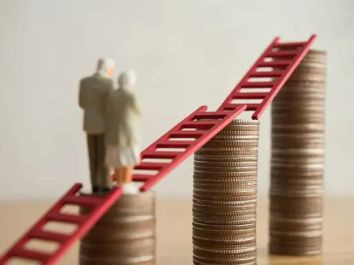 Figurki starszej pary stoją na stosach monet, połączonych czerwoną drabiną. Symbolizuje to nowe zasady przejścia na emeryturę i drogę do finansowej stabilności.
