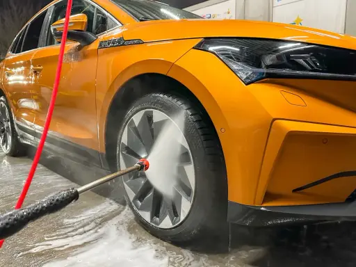 Pomarańczowe auto w trakcie mycia. Woda pod ciśnieniem spływa po karoserii i felgach, tworząc pianę.
