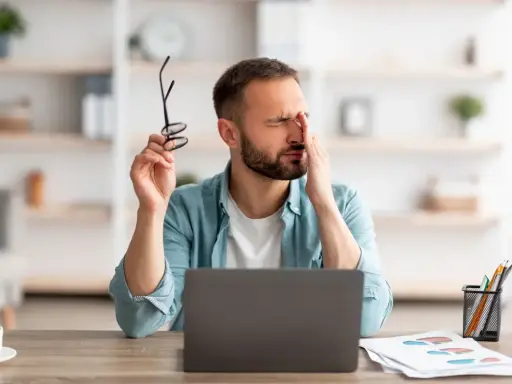 Homme fatigué, se frottant les yeux, lunettes à la main, devant son ordinateur. Il souffre de mal aux yeux à cause des écrans.