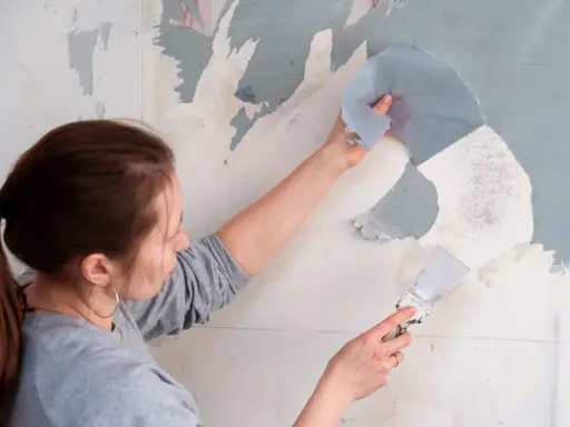 Une femme utilise une spatule pour décoller du papier peint ancien.
