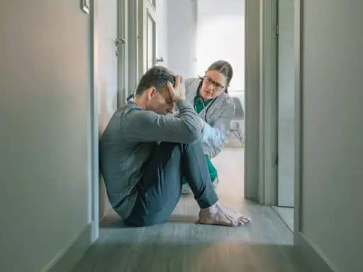 Mężczyzna w pozycji embrionalnej, z rękami na głowie, doświadcza objawów katatonii. Lekarka w białym fartuchu pochyla się nad nim, oferując wsparcie.