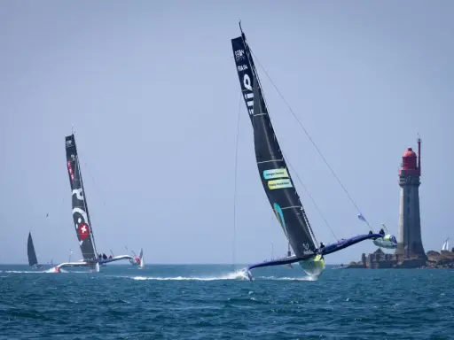 Course d'Ocean Fifty près d'un phare. Les trimarans filent sur l'eau, leurs voiles noires portant des logos.