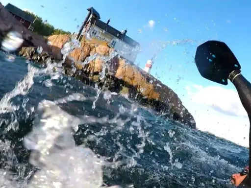 Vattenstänk och en paddel i förgrunden. I bakgrunden en klippa med hus och ett litet fyrhus. En del av en farleden swimrun-deltagare syns.