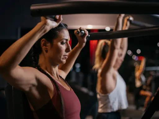 Zwei Frauen trainieren im Fitnessstudio. Sie nutzen eine Maschine, die zeigt, welche Muskelgruppen zusammen trainieren.