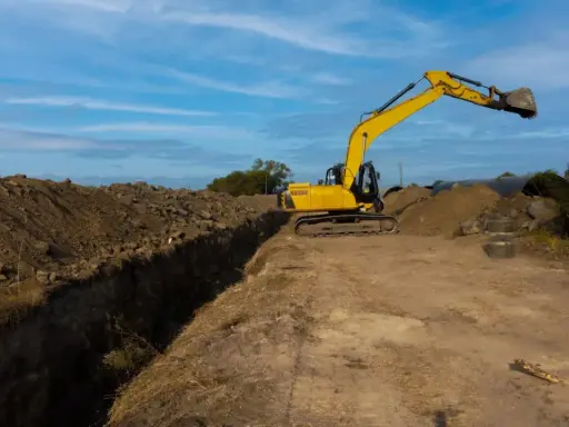 Excavadora amarilla trabajando en la creación de varios tipos de zanjas, con montones de tierra y tuberías grandes en el fondo.