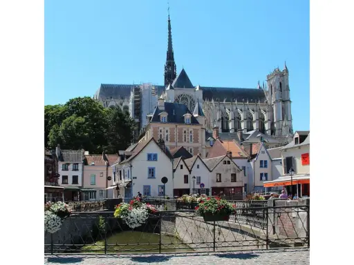 Blick auf malerische Häuser und die Kathedrale in Amiens, Frankreich. Blumenkästen schmücken eine Brücke über einen Kanal.