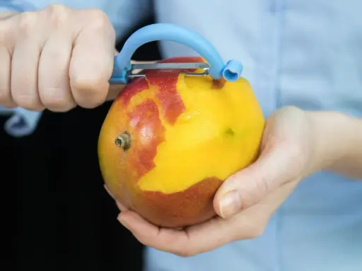 Hände schälen eine reife Mango mit einem blauen Sparschäler. Das Fruchtfleisch leuchtet gelb, während die Schale rot und gelb ist.