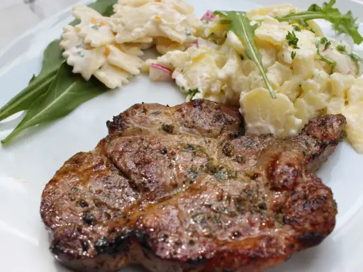 Saftiges Nackensteak grillen mit Kartoffel- und Nudelsalat auf einem weißen Teller.