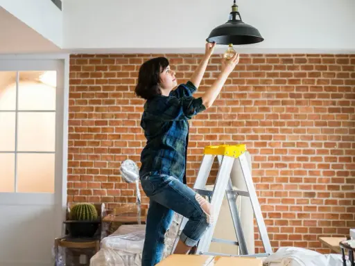 Une femme sur une échelle change une ampoule. L'indemnisation assurance obligation faire travaux est en cours.