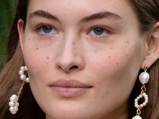 Primer plano de un rostro femenino con pecas naturales salpicando su piel. Lleva pendientes de perlas colgantes.