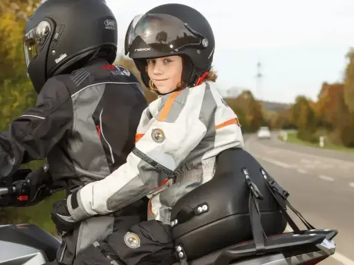 Uśmiechnięte dziecko na motocyklu, w kasku i kurtce, siedzi za kierowcą.