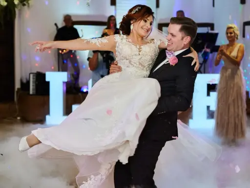 Pan młody niesie pannę młodą, tańcząc przy świetlnych literach LOVE. Idealny moment, by zastanowić się, co zaśpiewać na weselu dla pary młodej.